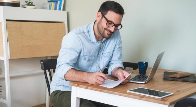 Cómo darte de alta como autónomo en 2026: trámites y obligaciones Cómo darte de alta como autónomo en 2026: trámites y obligaciones. Hombre sonriente sentado en una mesa con computadora portátil y tableta, escribiendo en papel foto de archivo