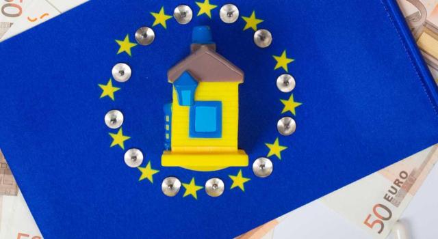 La Comisión Europea insta a España a que ponga fin a la imposición discriminatoria de las viviendas de contribuyentes no residentes que trabajan y residen habitualmente en España. Imagen de una casa amarilla rodeada de la bandera de la UE y billetes de 50 euros