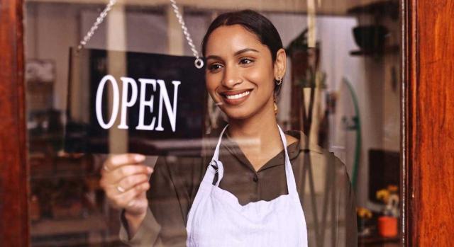 Reducción inicio de actividad: pasar de estimación objetiva a estimación directa Iniciar la actividad económica bajo el régimen de estimación objetiva, impide aplicar la reducción por inicio de actividad el siguiente año que se pase a estimación directa. Imagen de una chica joven muy sonriendo poniendo el cartel de open en una tienda