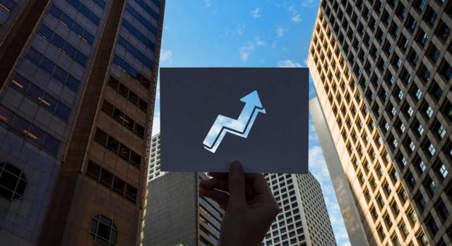 El pago contingente asociado a ciertas variables se considera mayor precio de adquisición de la edificación y su amortización será deducible en IS cuando responda a su depreciación efectiva. Imagen de una mano con una gráfica entre rascacielos