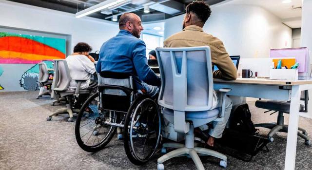 Se aplica la reducción en el IRPF para trabajadores con discapacidad aunque no exista habitualidad en la prestación laboral. Dos compañeros de trabajo hablando, uno de ellos en silla de ruedas en una oficina