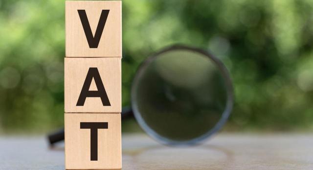 Ley  11/2021: modificaciones en IVA e IGIC. Imagen de cubos de madera superpuestos verticalmente con la palabra VAT escrita y sobre fondo difuminado con lupa
